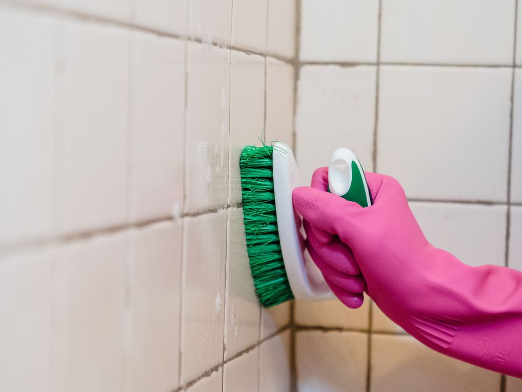 Gloved hand scrubbing discoloured grout lines between white wall tiles with a stiff brush